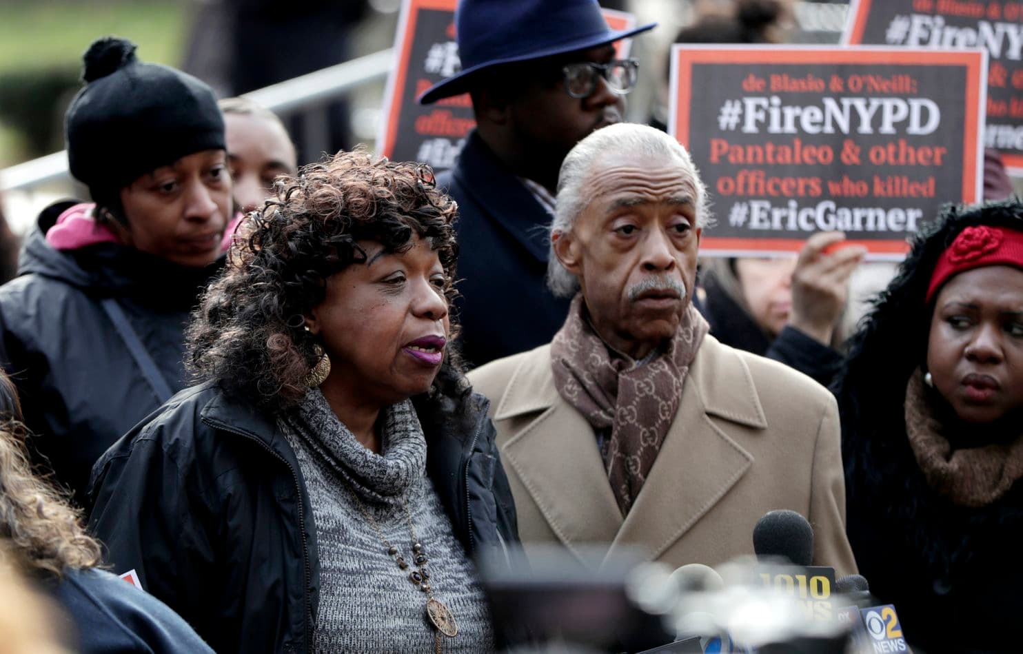 Rev. Sharpton and Gwen Carr, mother of Eric Garner, at press conference following disciplinary hearing for Eric Garner's killer, with signs denouncing NYPD in background.